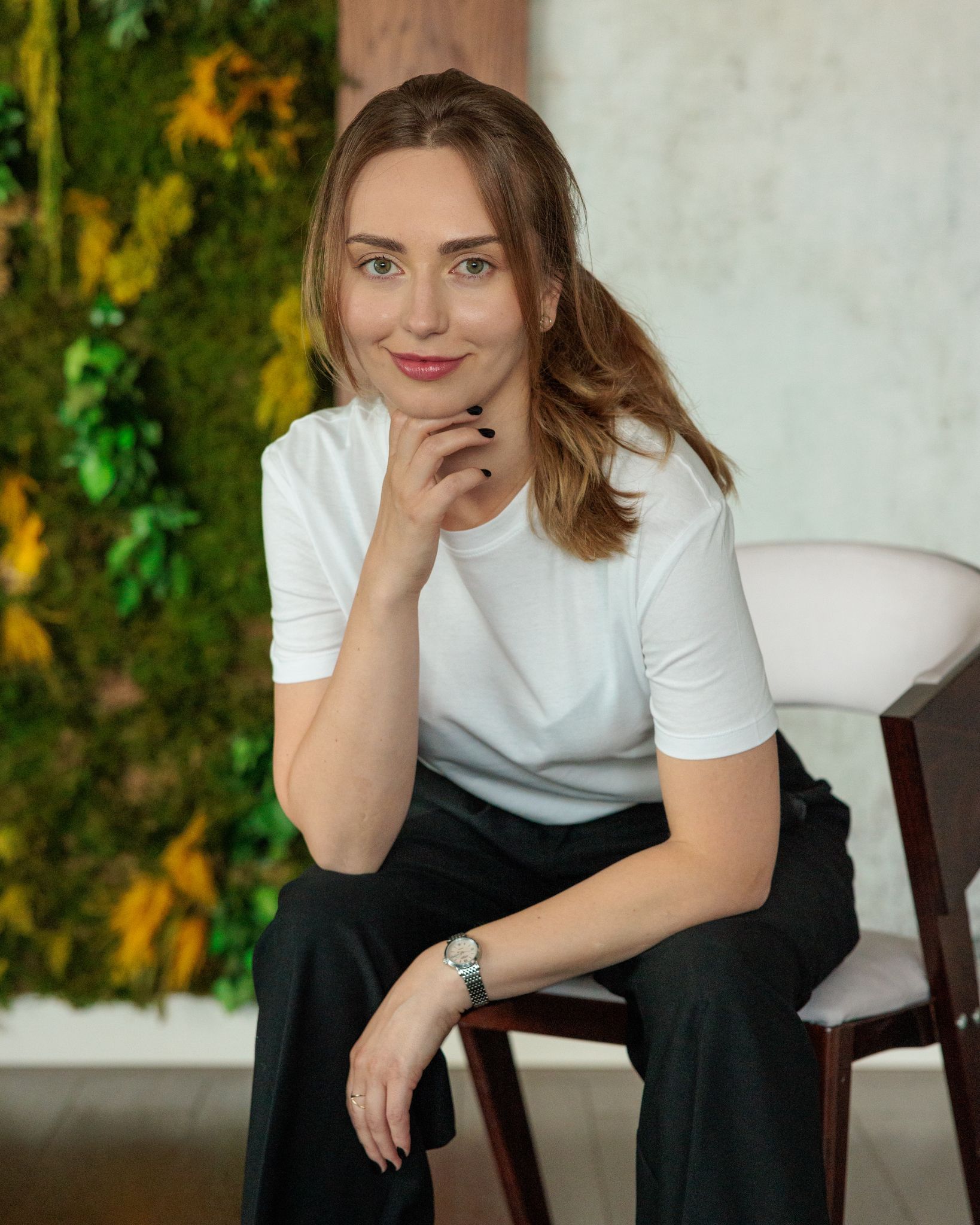 Anastasia Goluk w białej koszulce siedzi na tle zielonej ściany z mchu i uśmiecha się ciepło